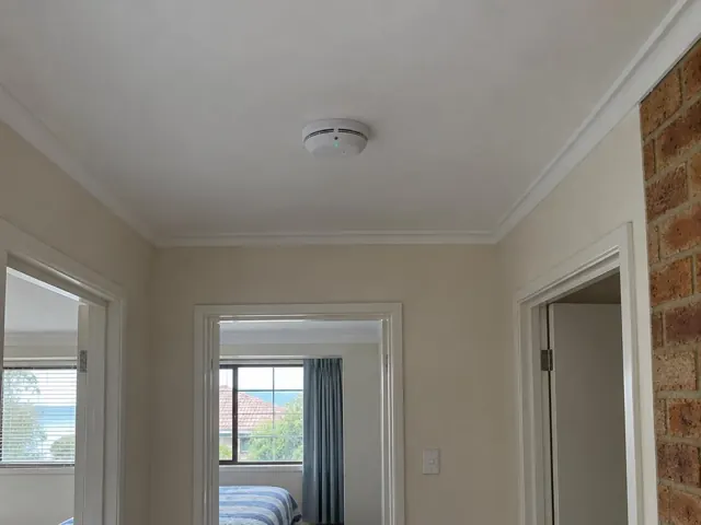 Ceiling smoke alarm in a beige hallway opening to a bedroom and brick wall in Booker Bay